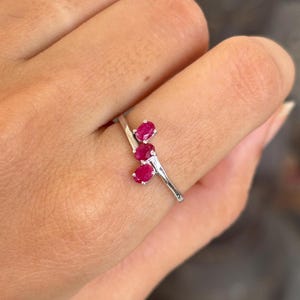 Op de afbeelding: Een zilveren ring met drie ovale, helderrode edelstenen. De edelstenen zijn in een cluster gerangschikt, wat een uniek en elegant ontwerp creëert. De ring wordt op een vinger getoond, wat de delicate en verfijnde uitstraling benadrukt.