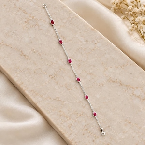 May include: A delicate silver chain bracelet featuring six evenly spaced, oval-shaped red gemstones set in smooth metal bezels. The bracelet includes a small circular clasp at one end and a matching jump ring at the other, showcasing a minimalist design.