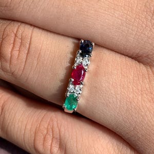 Può includere: Un anello d'argento con una fila di pietre preziose. L'anello presenta uno smeraldo verde, un rubino rosso, un diamante bianco e uno zaffiro blu.