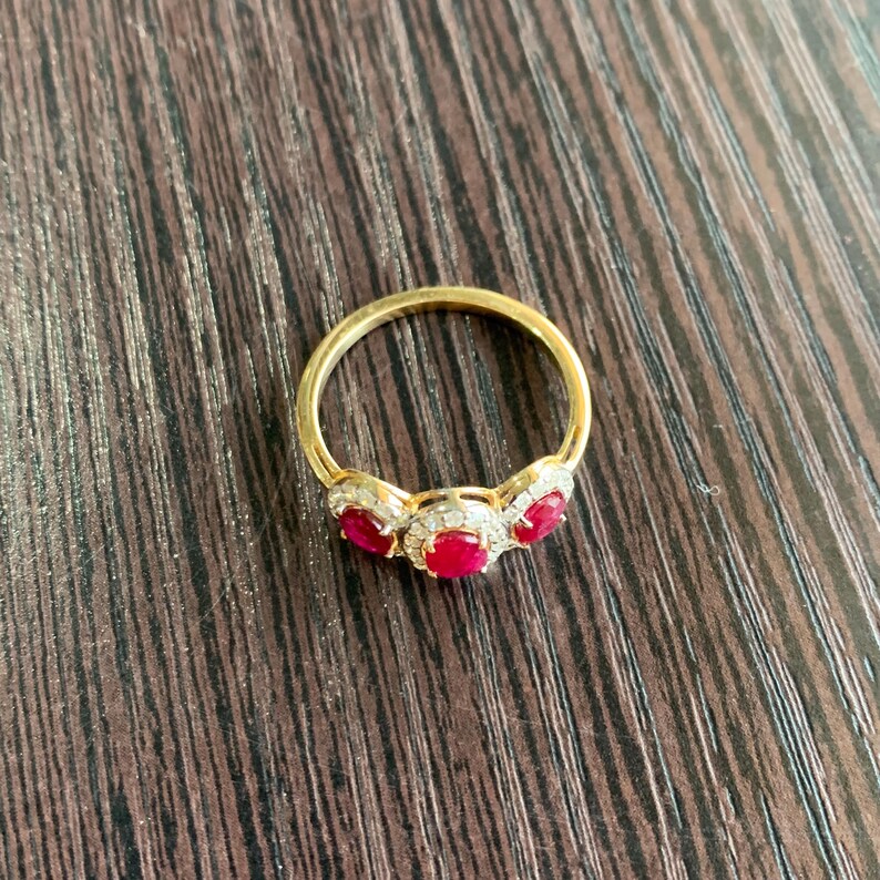 May include: A gold ring featuring three oval-cut, vibrant red gemstones, each surrounded by a halo of smaller, clear stones. The ring's band is a simple, polished gold. The ring is displayed on a dark wood surface.