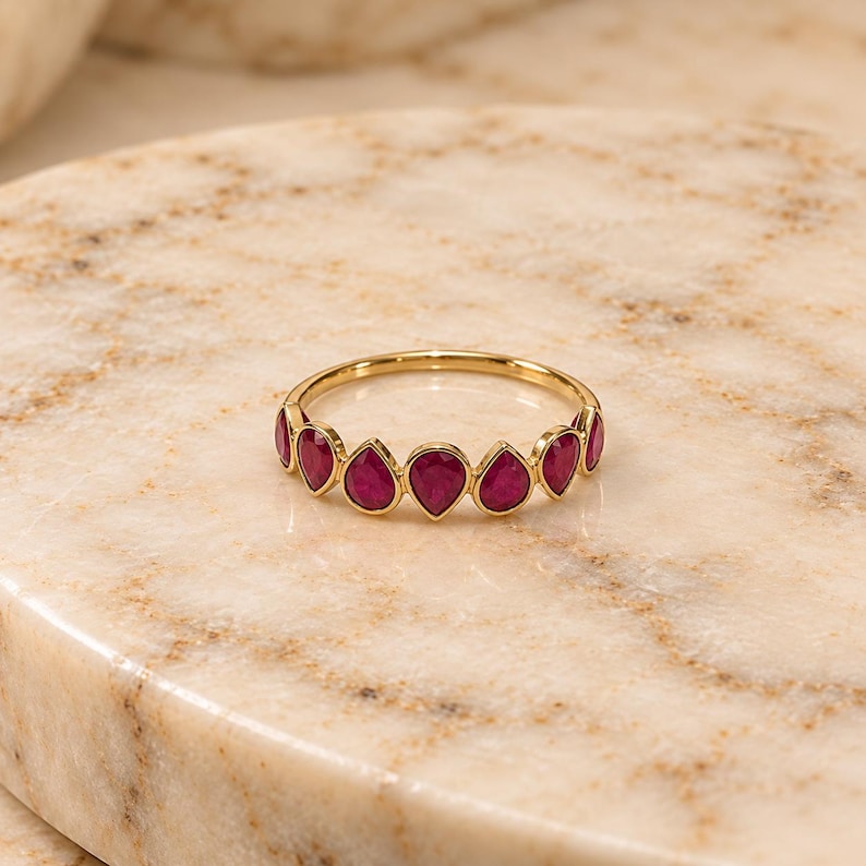 May include: A gold ring featuring a series of seven teardrop-shaped, deep red gemstones set in a row along the band. The stones are arranged in a bezel setting, creating a continuous decorative arc across the front of the thin metal band.