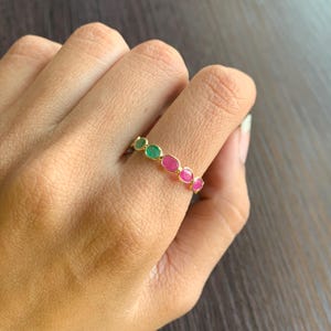 May include: A gold ring featuring a series of alternating green and pink gemstones. The gemstones are oval-shaped and set in a gold band. The ring is shown on a person's finger.