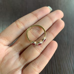 May include: A gold ring featuring a series of alternating red gemstones and small white diamonds. The ring is held in a hand, showcasing the intricate design and sparkle of the jewelry. The gemstones are set in a delicate, elegant arrangement.