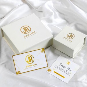 以下が含まれることがあります： 「Jewels Amo You desire we deliver」と金色の文字で書かれた白い箱が2つと、同じロゴと「Thank You For choosing Jewels Amo, where every piece is crafted with elegance and passion just for you www.jewelsamo.com」というテキストが書かれたサンキューカード。
