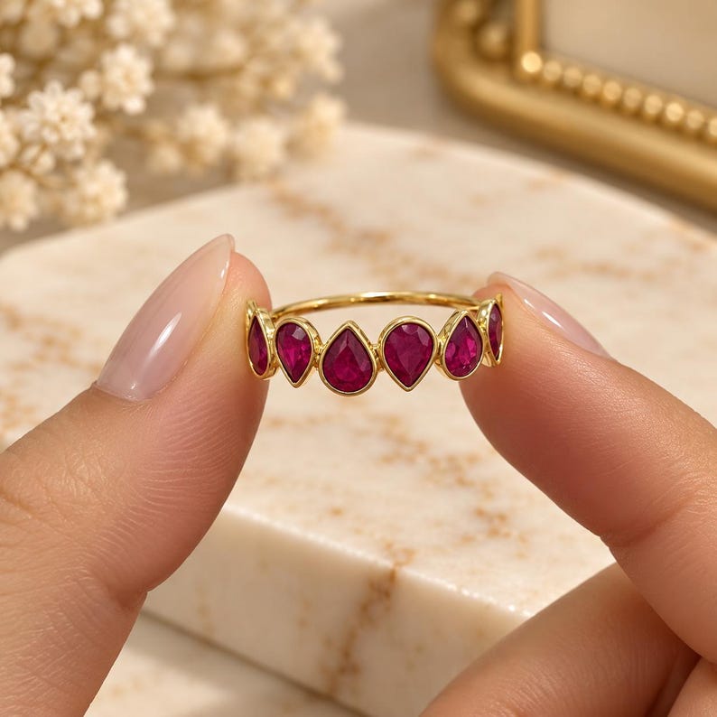 May include: A gold ring featuring a series of bezel-set, pear-shaped, deep pinkish-red gemstones arranged in a row along the band. The metal band is thin and polished, highlighting the vibrant color and teardrop cut of the stones.