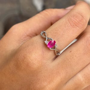 May include: A silver ring with a vivid oval ruby gemstone at its centre. The ring has an intricate, interwoven design, with small accent stones either side of the ruby, adding sparkle and detail to the jewellery.