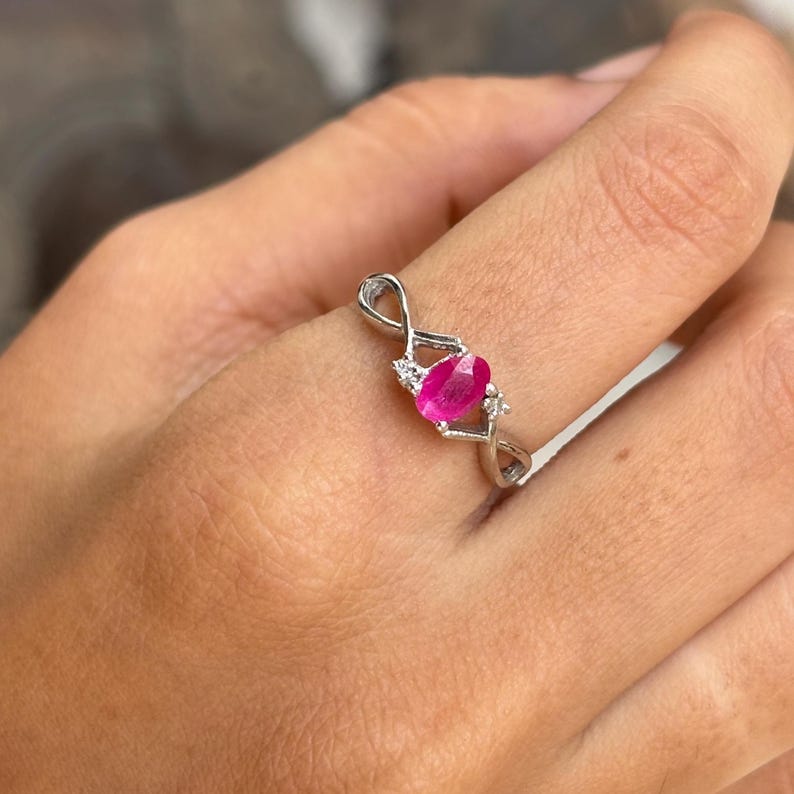 May include: A silver ring with a vivid oval ruby gemstone at its centre. The ring has an intricate, interwoven design, with small accent stones either side of the ruby, adding sparkle and detail to the jewellery.