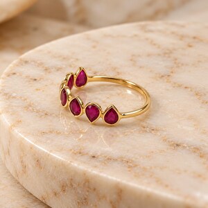 May include: A gold ring featuring a series of five teardrop-shaped, deep pink rubies set along the top half of the band. The stones are arranged in a row, each secured in a smooth gold bezel setting, creating a continuous, delicate design.