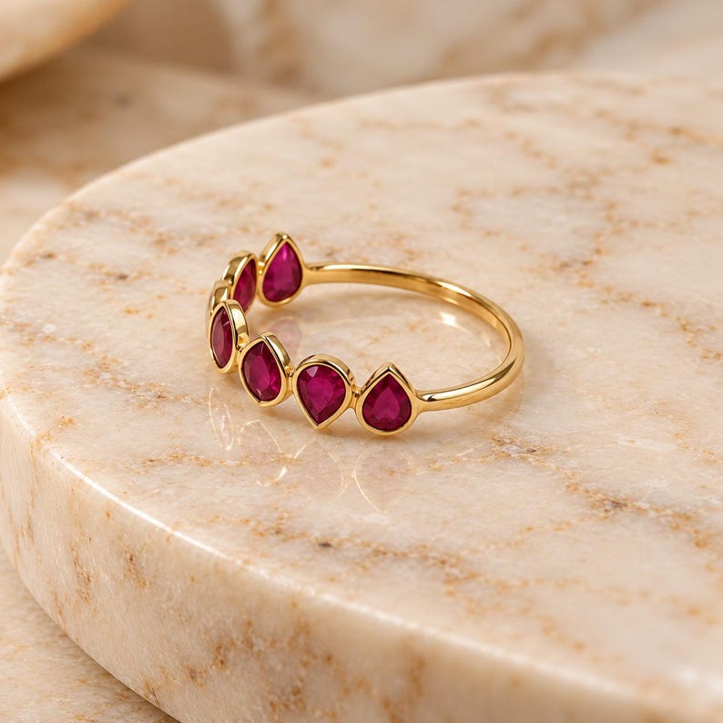 May include: A gold ring featuring a series of five teardrop-shaped, deep pink rubies set along the top half of the band. The stones are arranged in a row, each secured in a smooth gold bezel setting, creating a continuous, delicate design.