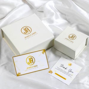 May include: White jewelry boxes and cards with gold accents are arranged on a white satin surface. The boxes and cards feature the logo "JA JEWELS AMO" in gold. One card reads "Thank You".