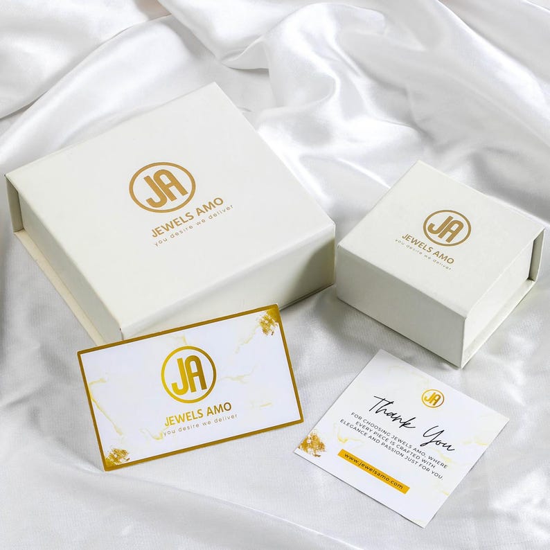 May include: White jewelry boxes and cards with gold accents are arranged on a white satin surface. The boxes and cards feature the logo "JA JEWELS AMO" in gold. One card reads "Thank You".