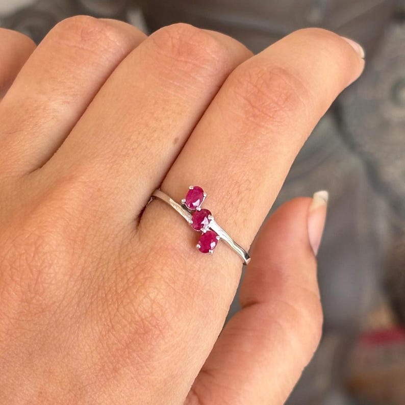 May include: A silver ring featuring three oval-cut, vibrant red gemstones. The gemstones are set in a cluster design on a thin, textured band. The ring is shown on a finger.