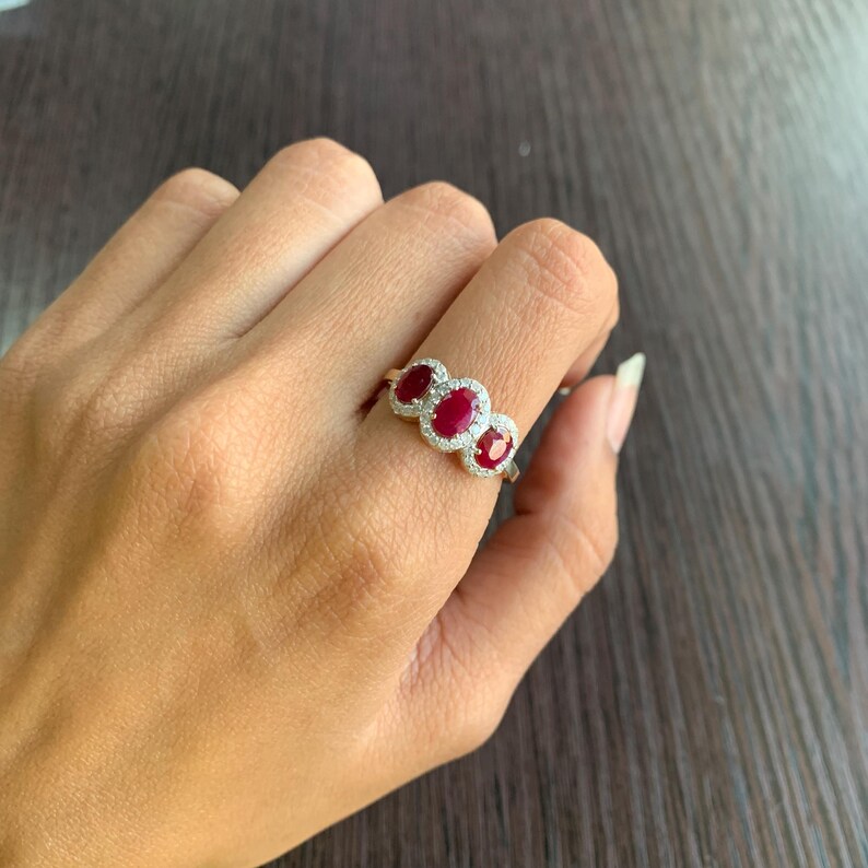 May include: A gold ring featuring three oval-cut, deep red gemstones, each surrounded by a halo of smaller, clear stones. The ring is set on a slender band, showcasing the vibrant red and sparkling accents.