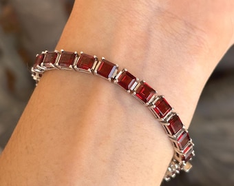 May include: A silver bracelet featuring rectangular, deep red gemstones. The gemstones are set in a row, creating a continuous pattern. The bracelet is shown on a wrist, highlighting the jewelry's design and color.