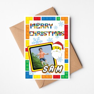 May include: A Christmas card with a colorful Lego-style border and the words "MERRY CHRISTMAS". It features a photo of a child jumping, a Santa Claus graphic, and the name "SAM". The card is on a brown envelope.