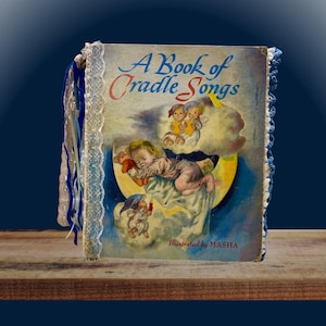 Op de afbeelding: Een vintage boek getiteld A Book of Cradle Songs met een illustratie van een slapende baby op een maansikkel, omringd door wolken en kleine figuren. Het boek heeft een witte kanten rand aan de linkerkant en een blauwe gehaakte rand aan de rechterkant.