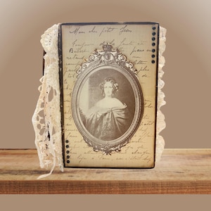 Op de afbeelding: Een rechthoekig dagboek met een crèmekleurige kaft voorzien van handgeschreven tekst, een centraal ovaal kader met een sepia-kleurig portret van een persoon, en decoratieve kanten randen aan de linker- en rechterkant van de rug.