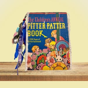 Op de afbeelding: Een vintage hardcover boek getiteld The Children's ANNUAL PITTER PATTER BOOK met 208 Pages of Entertainment. De blauwe kaft bevat kleurrijke illustraties van kinderen en een konijn, met decoratieve stoffen linten en kralen aan de rug bevestigd.