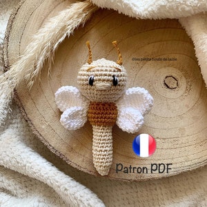 Può includere: Un sonaglio a forma di ape fatto a crochet con ali bianche e un corpo marrone e beige. Il giocattolo è su una superficie di legno con una coperta bianca e soffice sullo sfondo. Il testo "Patron PDF" è visibile nell'immagine.