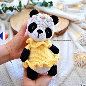 May include: A crocheted panda bear wearing a yellow dress with a ruffled hem and a floral headband. The panda is holding a small white heart in its paws.