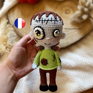 Peut inclure: Une poupée zombie en crochet avec des yeux verts, des cheveux bruns et une chemise verte. La poupée porte un pantalon marron et des chaussures noires. La poupée tient un petit objet brun dans sa main. © les petits bouts de laine