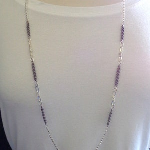 May include: A silver chain necklace with alternating silver and purple links. The necklace is on a white shirt with a patterned skirt.