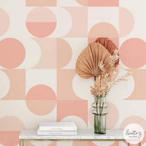 May include: A peach-coloured geometric wallpaper with a white background. The wallpaper features a repeating pattern of squares and half circles. A vase with dried flowers sits on a white marble table in front of the wallpaper.