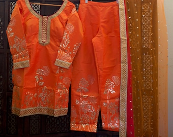 May include: An orange salwar kameez with silver floral print and gold trim. The outfit includes a long-sleeved top, pants, and a sheer dupatta with gold and red trim.