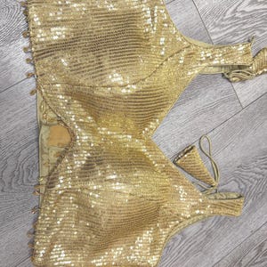May include: A sleeveless gold sequin crop top featuring a deep V-neckline, structured cups, and a side zipper closure. The garment is adorned with small dangling gold bead accents along the left side and includes thin matching fabric ties.