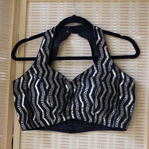 May include: A black halter top with a gold zigzag pattern, hanging on a black hanger. The top has a V-neck design and is made of a soft, possibly velvet-like material. The background is a light brown bamboo screen.