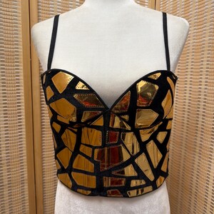 May include: A black bustier top with gold geometric shapes. The top has thin black straps and a sweetheart neckline. The gold shapes are arranged in a mosaic pattern across the front of the bustier.
