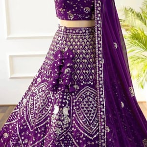 May include: A vibrant purple formal dress with intricate silver embellishments. The dress features a crop top, a full skirt, and a matching sheer scarf. The design includes detailed patterns and a ruffled accent on the skirt, creating an elegant look.
