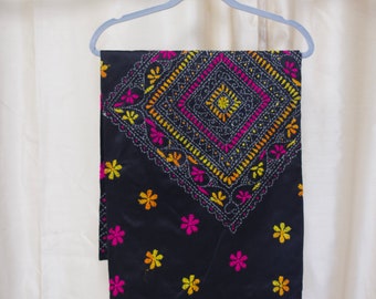 May include: A black fabric with colorful embroidery. The fabric has a diamond pattern with floral accents in pink, yellow, and orange. The fabric is hanging on a hanger.