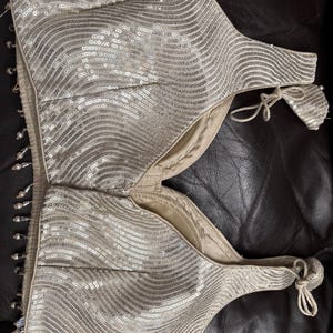 May include: A silver sequined top with a wavy pattern. The top has a scoop neckline and ties at the shoulders. The edges are trimmed with small, dangling beads. The garment is made of a shiny, reflective material.