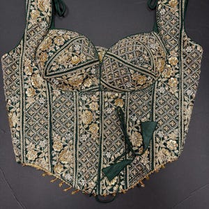 May include: A dark green corset top featuring intricate gold embroidery, floral patterns, and geometric beadwork. The garment has a square neckline, structured cups, wide shoulder straps, and a tie-front closure with dangling gold-toned accents.