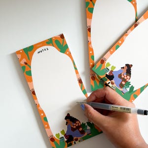 Könnte beinhalten: Weißer Notizblock mit einem orangefarbenen und grünen Blumenrand. Das Wort "NOTES" ist oben aufgedruckt. Der Notizblock zeigt eine Illustration einer Person, die gärtnert. Eine Hand hält einen Stift und schreibt auf den Notizblock.