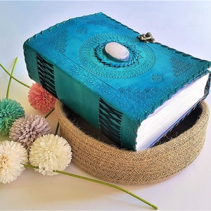 Peut inclure: Un journal en cuir turquoise avec un motif en relief et une pierre ovale au centre. Le journal est fermé par un fermoir en métal et présente une reliure cousue. Il repose dans un panier tressé, avec des fleurs décoratives à proximité.