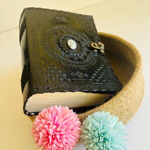 May include: A black leather journal with a decorative design and a silver clasp. The journal is sitting in a woven basket with two pink and blue flowers.