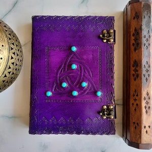 May include: Purple leather journal with a Celtic knot design and turquoise gemstones. The journal has two brass clasps and a decorative border.