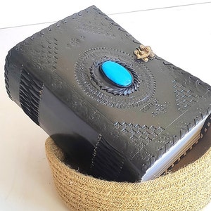 May include: A black leather-bound journal with intricate embossed designs and a turquoise gemstone set in a metal clasp. The journal is resting on a woven jute basket.