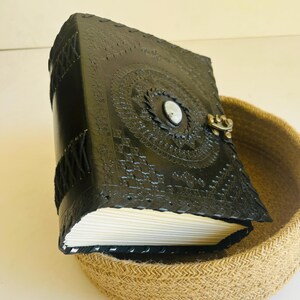 May include: A black leather journal with intricate embossed designs and a white stone closure. The journal is sitting in a woven basket.