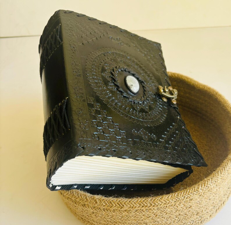 May include: A black leather journal with intricate embossed designs and a white stone closure. The journal is sitting in a woven basket.