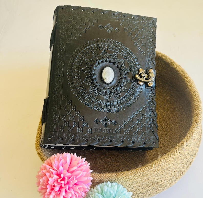 May include: Black leather journal with intricate embossed designs and a white stone accent. The journal has a clasp closure and is sitting on a woven basket.