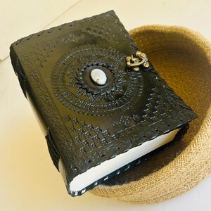 May include: Black leather journal with intricate embossed designs and a silver metal clasp. The journal is closed and sitting on a woven basket.