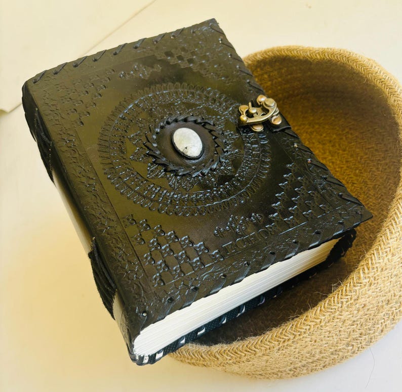 May include: Black leather journal with intricate embossed designs and a silver metal clasp. The journal is closed and sitting on a woven basket.