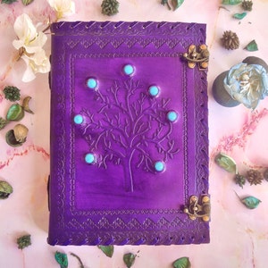 Puede incluir: Diario de cuero morado con un diseño de árbol en relieve en la portada. El árbol tiene piedras preciosas turquesa incrustadas en las ramas. El diario tiene dos cierres metálicos.