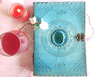 Pode incluir: Um diário de couro azul turquesa com um design intrincado e uma gema verde no centro. O diário tem um fecho de fivela e está rodeado por velas e uma flor.