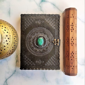 May include: A black leather journal with intricate embossed designs and a green gemstone closure. The journal is next to a wooden incense holder with a decorative pattern.