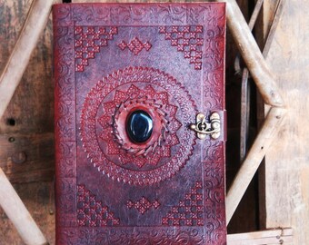 Pode incluir: Um diário de couro bordô com desenhos em relevo intrincados e um padrão circular de mandala. O diário tem um fecho de metal e uma pedra azul escura no centro do mandala.