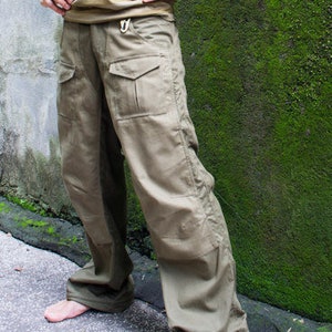 May include: A man wearing a striped tank top and khaki cargo pants stands in front of a mossy wall. He is looking up and has his hands on his hips.